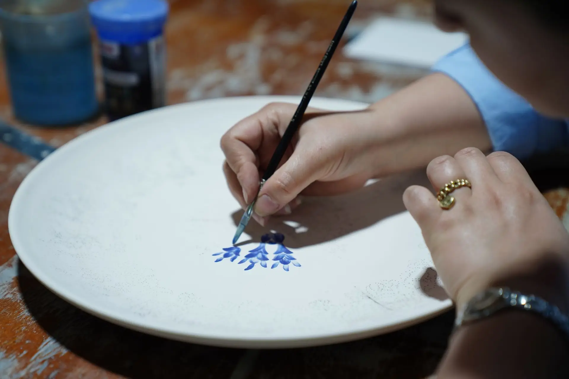Manos pintando flores azules con pincel sobre un plato blanco